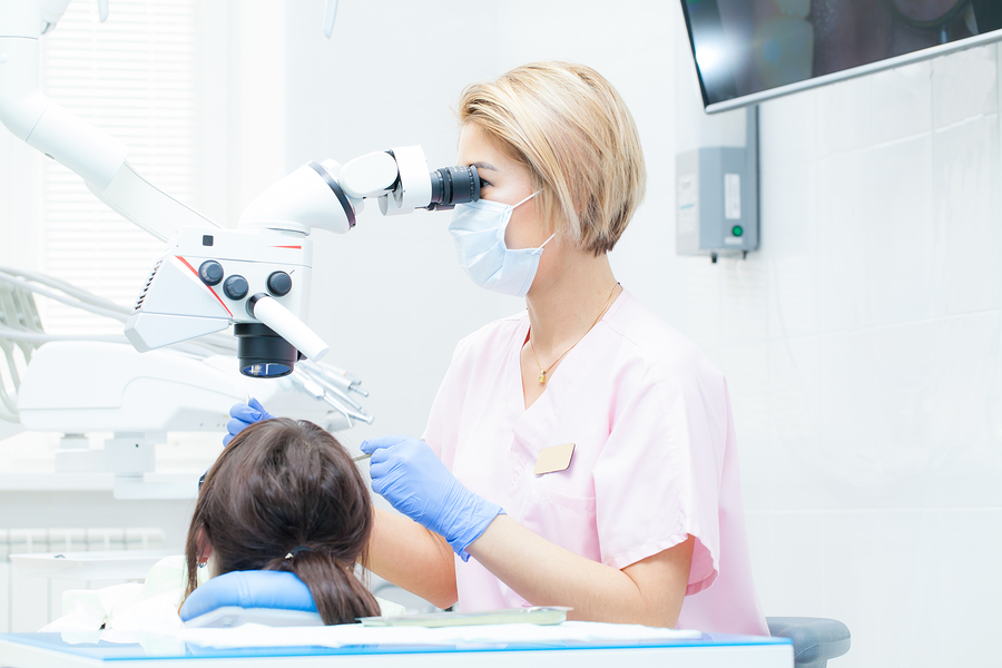Young female dentist treating root canals using microscope at the dental clinic. Young woman patient lying on dentist chair with open mouth. Dentist wearing mask and gloves