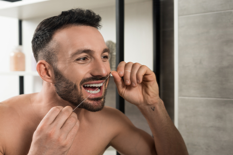bearded shirtless man looking at mirror while using dental floss in bathroom